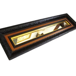 May include: A rectangular wall mirror featuring a long, narrow reflective glass surface. The frame consists of an outer black border with a carved leaf-like pattern, surrounding an inner border of warm brown wood and a thin gold-toned trim.