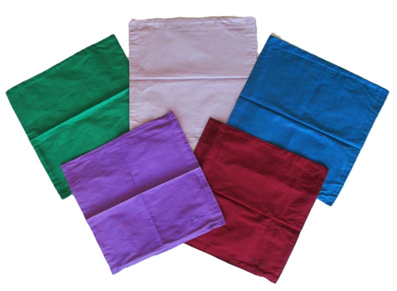 May include: A collection of five square fabric cushion covers arranged in a fan shape. The covers are solid colours including green, light pink, blue, purple, and dark red. Each square cover features a horizontal seam across the middle section.