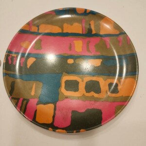 May include: A round decorative plate with a colorful abstract design. The plate features a mix of colors, including pink, orange, teal, and brown, arranged in a geometric pattern. The plate is likely made of ceramic or a similar material.