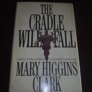 May include: A hardcover book titled "THE CRADLE WILL FALL" by Mary Higgins Clark. The cover features a dark illustration of a figure wrapped in a snake-like design. The book's title and author's name are in bold, black font.
