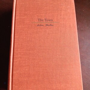 May include: A hardcover book titled "The Town" by William Faulkner. The book has a textured, reddish-orange cover with the title and author's name in a simple font. The book is closed and appears to be in good condition.