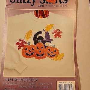 May include: A white long-sleeve shirt with a Halloween-themed iron-on applique kit. The design features a black cat wearing a witch hat, three orange jack-o'-lanterns, and colorful leaves. The package reads "Glitzy Shirts" and "SCAREDY-CAT".