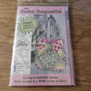 May include: A sewing pattern for "The Boho Baguette" purse, featuring a floral design in pink and green. The package includes the text "Flirty! Feminine & Fun to Carry" and purse dimensions of 12" x 12" x 7".