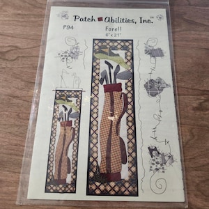 May include: A golf-themed fabric panel with the text "Fore!!" and dimensions of 6" x 21". The design features a golf bag with clubs, a golf cart, and decorative stitching. The color palette includes brown, beige, and green.