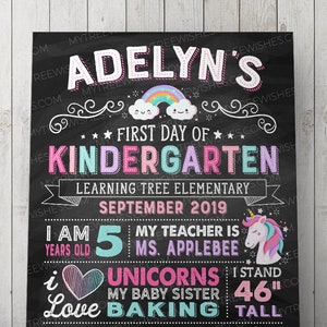 May include: A chalkboard-style sign with the text "Adelyn's First Day of Kindergarten." It includes a rainbow, unicorn, and various facts about the child, including her height of 46 inches. The sign is decorated with stars and swirls.