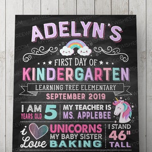 May include: A black chalkboard-style sign with colorful text and illustrations. The sign reads "Adelyn's First Day of Kindergarten" with a rainbow and cloud graphic. It includes details about the child's age, teacher, and favorite things. The child is 46 inches tall.