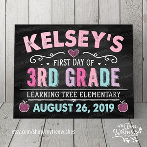 May include: A chalkboard-style sign with the text "Kelsey's First Day of 3rd Grade" in pink and blue lettering. The sign also includes the school name, date, and apple illustrations. The background is black.