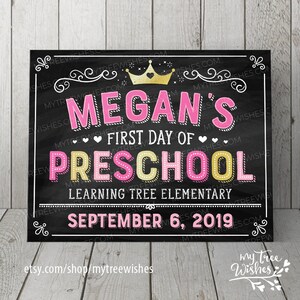 May include: A chalkboard-style sign with the text "Megan's First Day of Preschool" in pink and yellow lettering. A gold crown is at the top. The date "September 6, 2019" is at the bottom. The sign is framed in white.