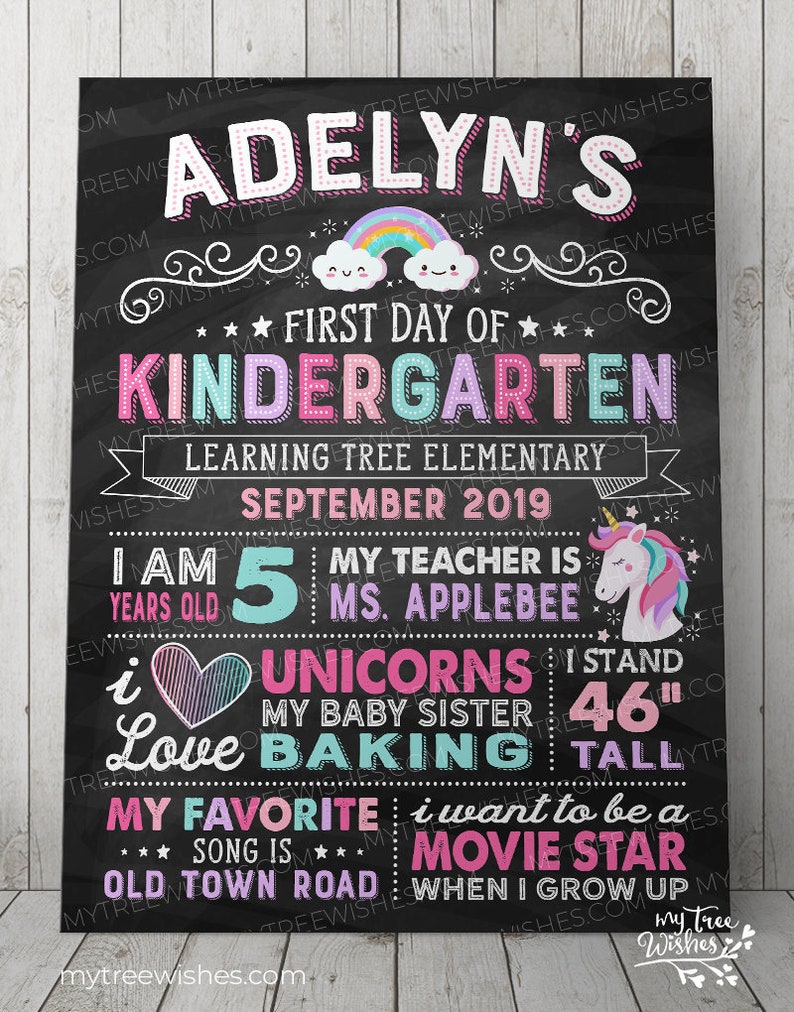 May include: A chalkboard-style sign with the text "Adelyn's First Day of Kindergarten." It includes a rainbow, unicorn, and various facts about the child, including her height of 46 inches. The sign is decorated with stars and swirls.