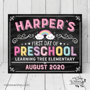May include: A chalkboard-style sign with the text "Harper's First Day of Preschool" in colorful letters. A rainbow and smiling clouds are featured above the word "Preschool". The sign also includes the text "Learning Tree Elementary" and "August 2020".