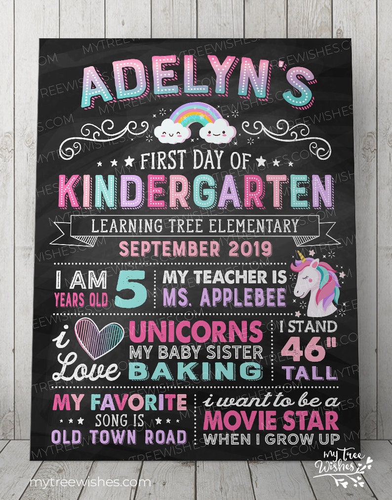 May include: A black chalkboard-style sign with colorful text and graphics. It reads "Adelyn's First Day of Kindergarten" with a rainbow and cloud graphic. Includes details about the child, including height of 46 inches, age, and favorite things.
