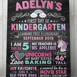 May include: A chalkboard-style sign for a child's first day of kindergarten. The sign features the name "Adelyn's" in pink, a rainbow, and the text "First Day of Kindergarten." It includes the child's age, teacher's name, and height of 46 inches.