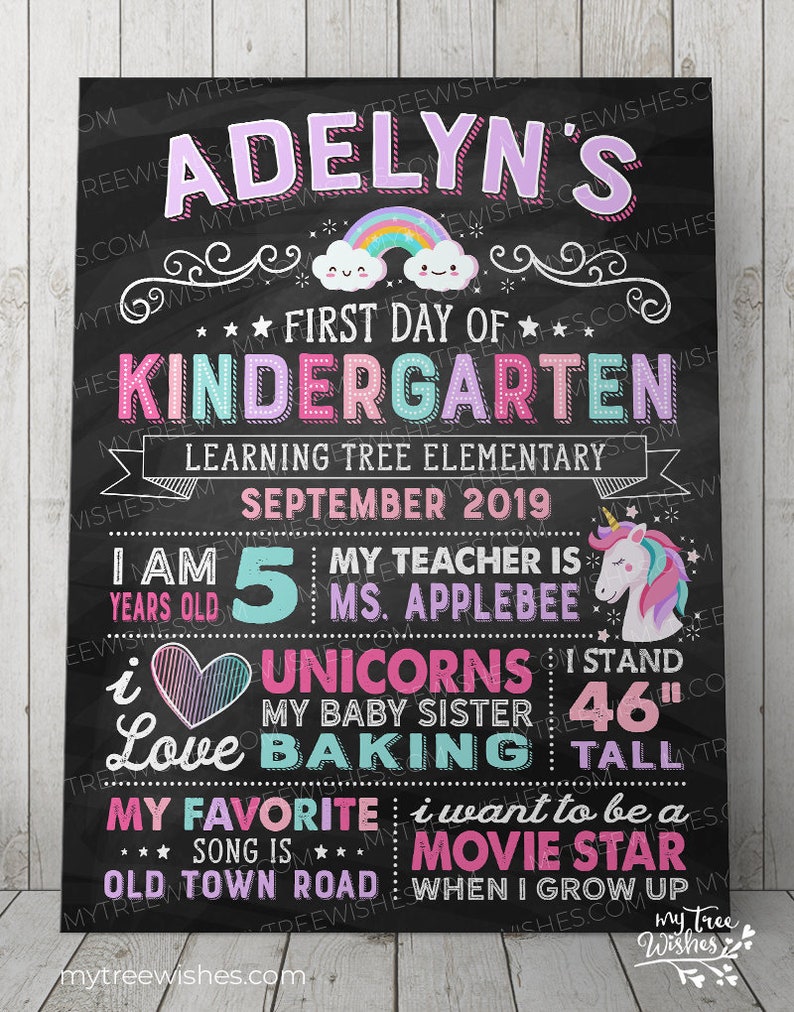May include: A black chalkboard-style sign with colorful text and illustrations. The sign reads "Adelyn's First Day of Kindergarten" with a rainbow and cloud graphic. It includes details about the child's age, teacher, and favorite things. The child is 46 inches tall.