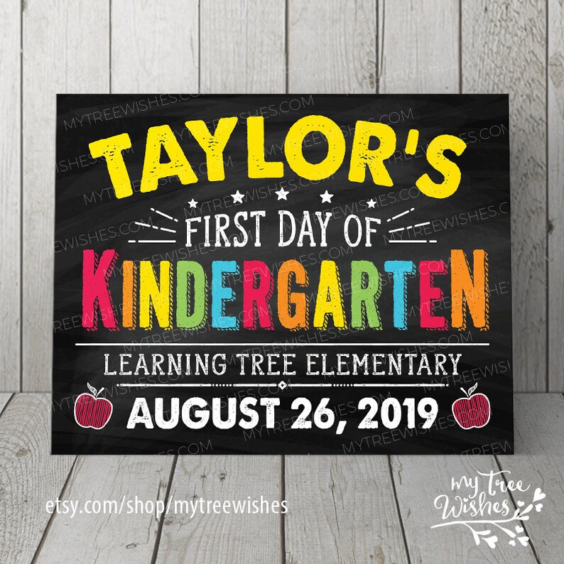 May include: A chalkboard-style sign with the text "Taylor's First Day of Kindergarten" in yellow, red, orange, green, and blue. It includes the school name, date, and apple illustrations. The sign is on a wooden surface.