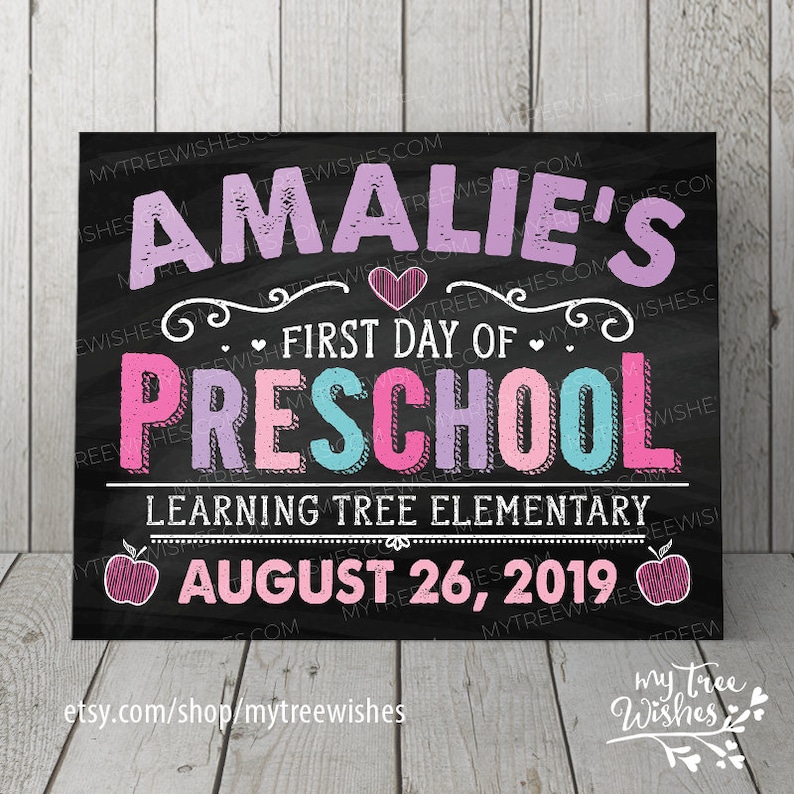 May include: A chalkboard-style sign with the text "Amalie's First Day of Preschool" in pink, purple, and teal lettering. The sign also includes the date August 26, 2019, and the name of the school, Learning Tree Elementary.