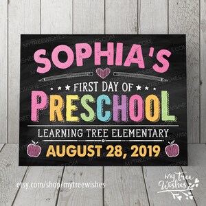 May include: A chalkboard-style sign with the text "SOPHIA'S FIRST DAY OF PRESCHOOL" in colorful letters. The sign includes the school name, date, and apple illustrations. The background is black with a wood-grain texture.