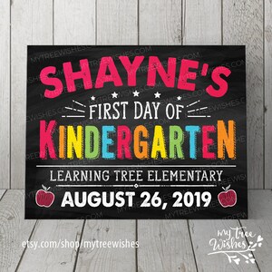 May include: A black chalkboard-style sign with the text "SHAYNE'S FIRST DAY OF KINDERGARTEN" in colorful letters. Below, it reads "LEARNING TREE ELEMENTARY AUGUST 26, 2019". Two red apple illustrations are on the sign.