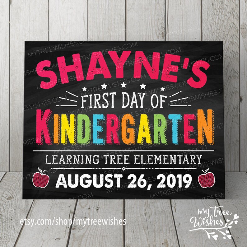 May include: A black chalkboard-style sign with the text "SHAYNE'S FIRST DAY OF KINDERGARTEN" in colorful letters. Below, it reads "LEARNING TREE ELEMENTARY AUGUST 26, 2019". Two red apple illustrations are on the sign.