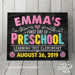 May include: A chalkboard-style sign with the text "EMMA'S FIRST DAY OF PRESCHOOL" in pink, yellow, and blue. The sign also includes the date "AUGUST 26, 2019" and the name of the school. The background is black.