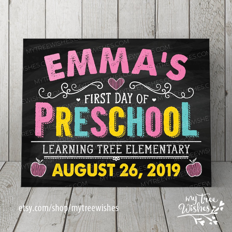 May include: A chalkboard-style sign with the text "EMMA'S FIRST DAY OF PRESCHOOL" in pink, yellow, and blue. The sign also includes the date "AUGUST 26, 2019" and the name of the school. The background is black.
