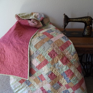 Op de afbeelding: Een patchworkdeken met een roze achterkant en een bloemenpatroon. De deken is gevouwen en gedrapeerd over een tafel met een vintage naaimachine erop.