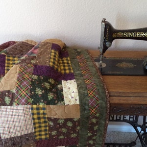 Peut inclure: Une machine à coudre Singer vintage avec une base en bois et une courtepointe en patchwork de tissus bruns, verts et violets.