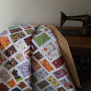 Könnte beinhalten: Eine bunte Patchwork-Decke mit weißem Hintergrund und verschiedenen Blumenmustern. Die Decke ist gefaltet und über einen Holztisch mit einer antiken Nähmaschine darauf gelegt. Die Nähmaschine ist schwarz mit goldenen Verzierungen und das Wort "SINGER" ist an der Seite sichtbar.