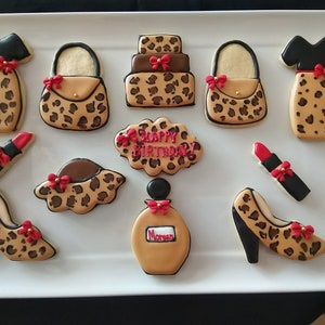 Puede incluir: Un conjunto de nueve galletas de azúcar decoradas con un diseño de estampado de leopardo. Las galletas tienen forma de vestido, bolso, tacones altos, lápiz labial, frasco de perfume y pastel. Una galleta dice "¡Feliz cumpleaños!" en glaseado marrón.