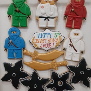 May include: A set of 10 decorated sugar cookies featuring a ninja theme. The cookies are decorated with green, blue, red, and white icing. The cookies include ninjas, swords, and throwing stars. The cookies are arranged on a white surface. The text "HAPPY 5th BIRTHDAY JACOB!" is written on one of the cookies.
