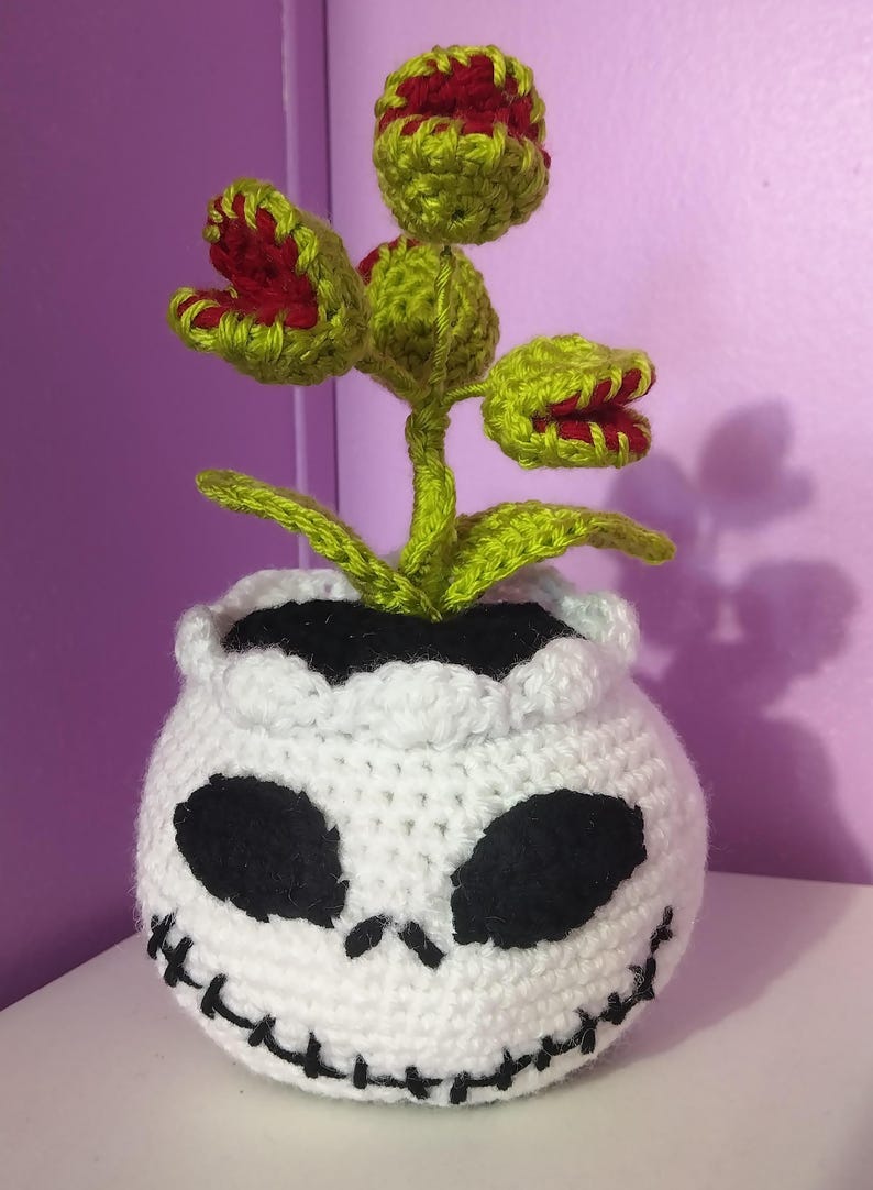 Puede incluir: Una planta tejida a ganchillo con tres cabezas de venus atrapamoscas verdes y rojas que emergen de una maceta blanca redonda con forma de calavera. La maceta presenta cuencas oculares negras bordadas y una boca cosida, con tallos y hojas verde brillante.