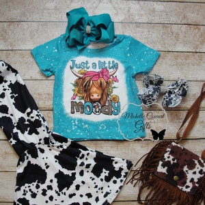 Pode incluir: Um conjunto infantil com uma camiseta turquesa com a frase "Just a little moody" e um gr&aacute;fico de vaca, combinado com leggings, sapatos, uma bolsa e um la&ccedil;o azul-petr&oacute;leo com estampa de vaca.