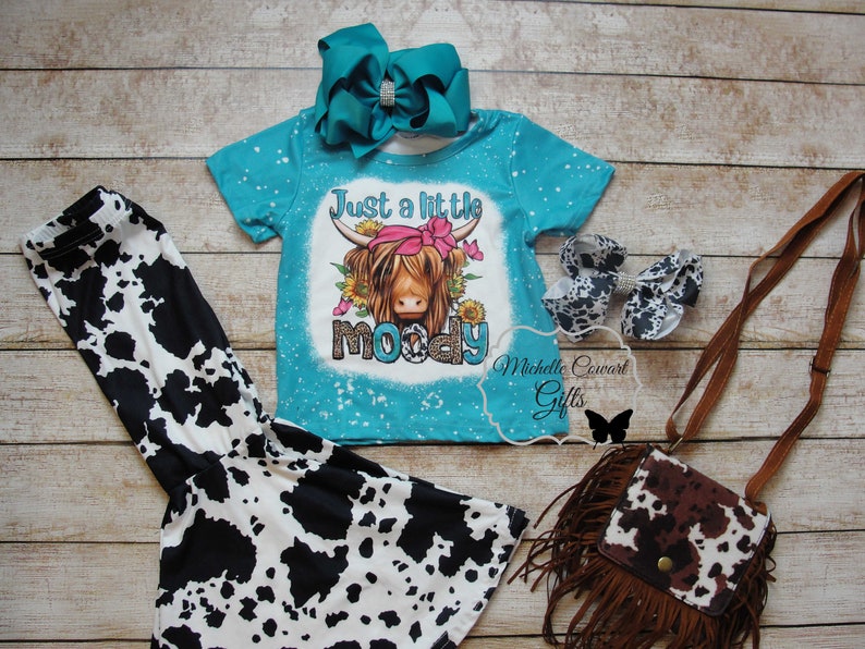Pode incluir: Um conjunto infantil com uma camiseta turquesa com a frase "Just a little moody" e um gr&aacute;fico de vaca, combinado com leggings, sapatos, uma bolsa e um la&ccedil;o azul-petr&oacute;leo com estampa de vaca.