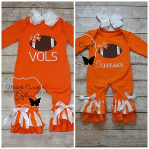 May include: Two orange baby onesies with white and orange striped ruffles. One onesie has a brown football with a white outline and the text "VOLS". The other onesie has a brown football with a white outline and the text "Tennessee".