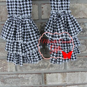 May include: Close-up of two black and white houndstooth patterned pants legs with tiered ruffles. The ruffles are trimmed with white lace. The pants are displayed against a brick wall. The text "Michelle Cornett Gifts" is visible.