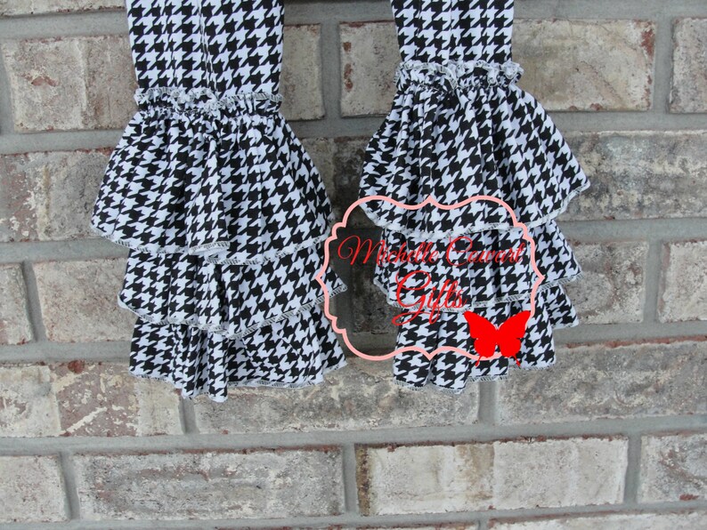 May include: Close-up of two black and white houndstooth patterned pants legs with tiered ruffles. The ruffles are trimmed with white lace. The pants are displayed against a brick wall. The text "Michelle Cornett Gifts" is visible.