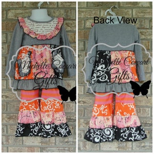 May include: A two-piece children's outfit featuring a long-sleeved gray top with a ruffled collar and floral print accents. The pants are tiered with orange, pink, and black floral patterns. The outfit is displayed on a mannequin.