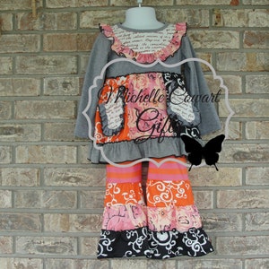 May include: A gray long-sleeved top and tiered pants set for a child. The top has a ruffled collar with floral and script details. The pants feature orange, pink, and black patterned tiers. The set is displayed on a mannequin against a brick wall.