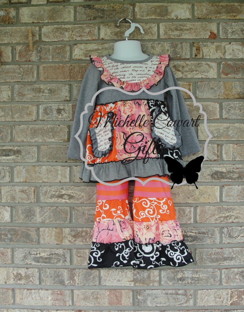 May include: A gray long-sleeved top and tiered pants set for a child. The top has a ruffled collar with floral and script details. The pants feature orange, pink, and black patterned tiers. The set is displayed on a mannequin against a brick wall.