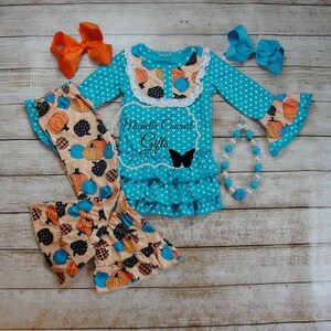 Puede incluir: Un conjunto turquesa con lunares con pantalones estampados de calabaza, una blusa de manga larga y lazos a juego. La blusa tiene un cuello de encaje y un dobladillo con volantes. Tambi&eacute;n se incluye un collar de cuentas con una rosa azul. El texto "Michelle Coward Gifts" es visible.