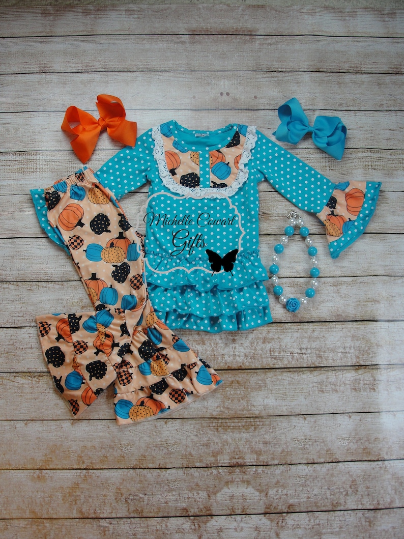 Puede incluir: Un conjunto turquesa con lunares con pantalones estampados de calabaza, una blusa de manga larga y lazos a juego. La blusa tiene un cuello de encaje y un dobladillo con volantes. Tambi&eacute;n se incluye un collar de cuentas con una rosa azul. El texto "Michelle Coward Gifts" es visible.