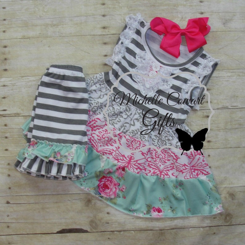 Pu&ograve; includere: Un abito senza maniche a righe grigie e bianche con una gonna a stampa floreale e un bordo in pizzo bianco. L'abito ha un fiocco rosa e una chiusura a bottone. L'abito &egrave; abbinato a leggings a righe grigie e bianche con un bordo a volant floreale. L'immagine include il testo "Michelle Cowart Gifts".