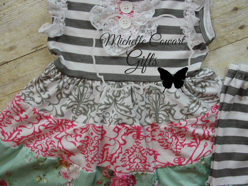 Pu&ograve; includere: Un vestito a righe grigie e bianche con un bordo di pizzo bianco e una gonna a strati con un motivo damascato grigio e bianco, un motivo floreale rosa e bianco e un motivo floreale verde e bianco. L'abito &egrave; abbinato a leggings a righe grigie e bianche.