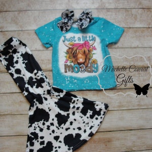 Pode incluir: Uma camiseta turquesa com um gr&aacute;fico de vaca e as palavras "Just a little moody". A camisa &eacute; combinada com cal&ccedil;as de sino com estampa de vaca em preto e branco e um la&ccedil;o combinando. Uma roupa divertida para crian&ccedil;as.