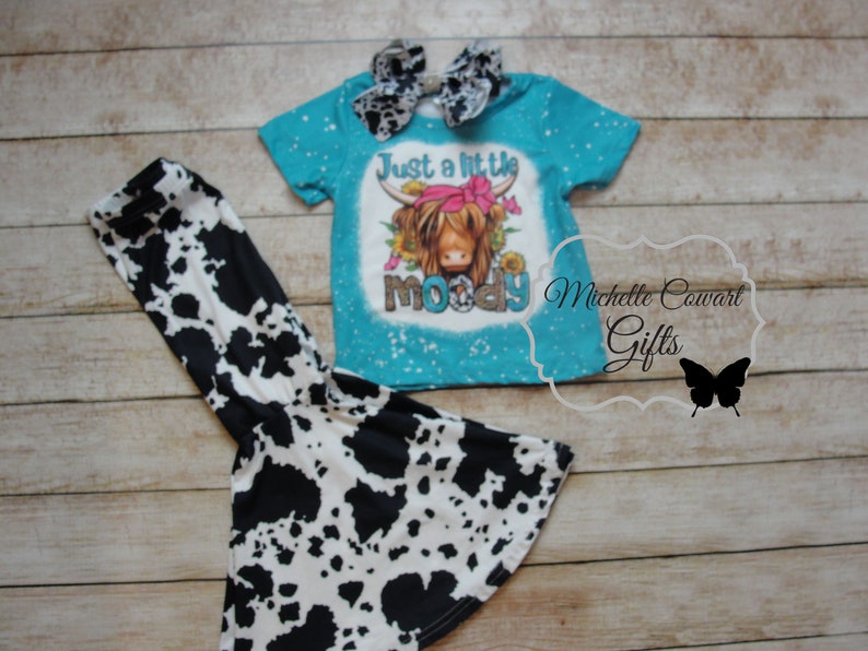 Pode incluir: Uma camiseta turquesa com um gr&aacute;fico de vaca e as palavras "Just a little moody". A camisa &eacute; combinada com cal&ccedil;as de sino com estampa de vaca em preto e branco e um la&ccedil;o combinando. Uma roupa divertida para crian&ccedil;as.