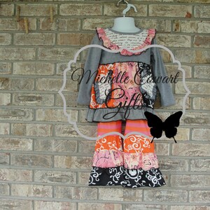 May include: A gray long-sleeved top and tiered pants set. The top has a ruffled collar with text and a floral design. The pants feature orange, pink, and black patterned tiers. The set is displayed on a hanger against a brick wall.