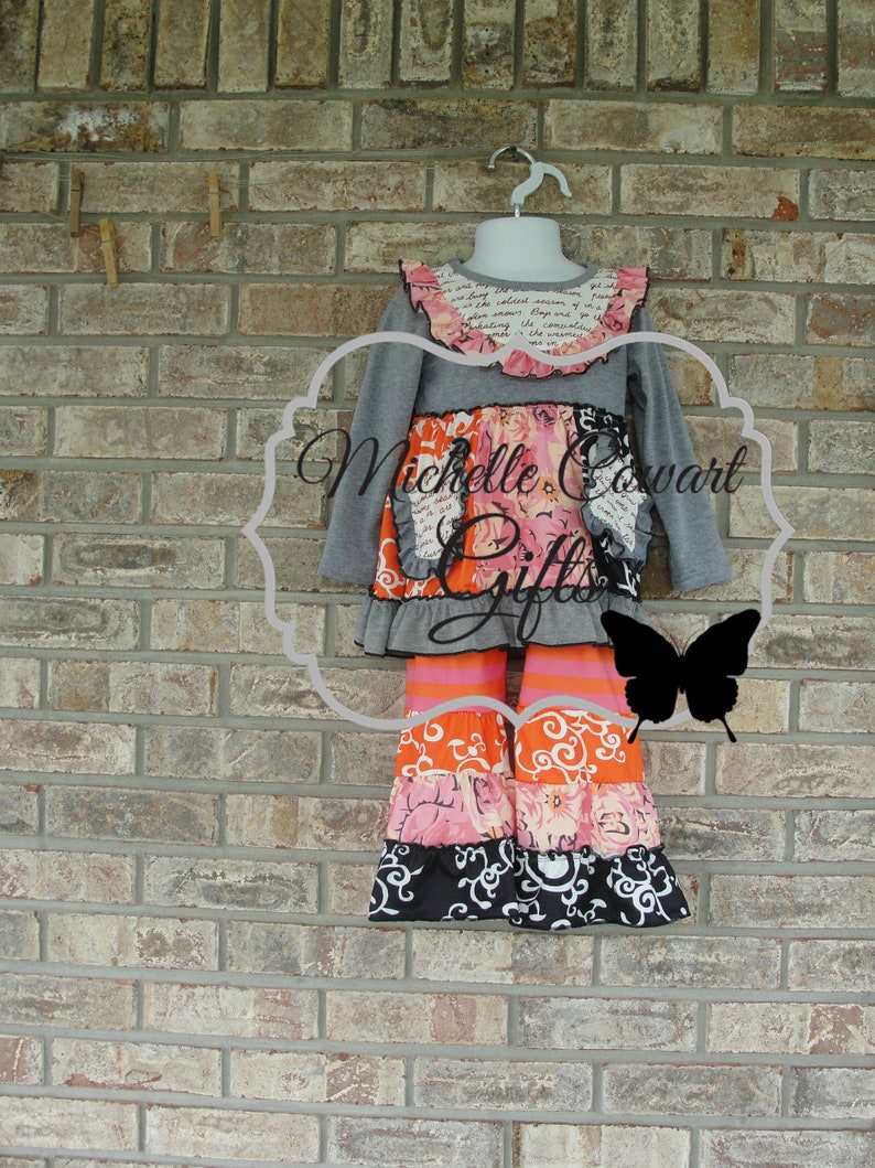 May include: A gray long-sleeved top and tiered pants set. The top has a ruffled collar with text and a floral design. The pants feature orange, pink, and black patterned tiers. The set is displayed on a hanger against a brick wall.