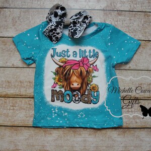 Pode incluir: Camiseta turquesa com um gr&aacute;fico de uma vaca Highland com um la&ccedil;o rosa e o texto "Just a little moody". A camisa tem manchas brancas e &eacute; combinada com um la&ccedil;o com estampa de vaca em preto e branco.