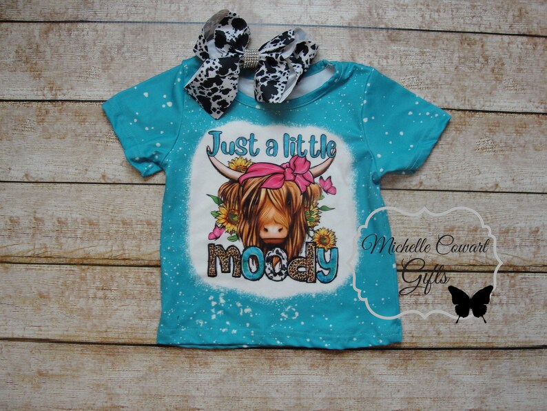 Pode incluir: Camiseta turquesa com um gr&aacute;fico de uma vaca Highland com um la&ccedil;o rosa e o texto "Just a little moody". A camisa tem manchas brancas e &eacute; combinada com um la&ccedil;o com estampa de vaca em preto e branco.