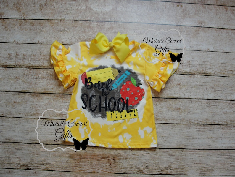 Op de afbeelding: Geel tie-dye shirt met een zwart-witte afbeelding met de tekst "Back to School" met een potlood, kleurpotlood en appel.