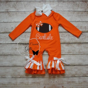 May include: Orange and white baby onesie with a brown football and white bow on the front. The onesie has a white ruffle trim and the name "Carlisle" is embroidered on the front.